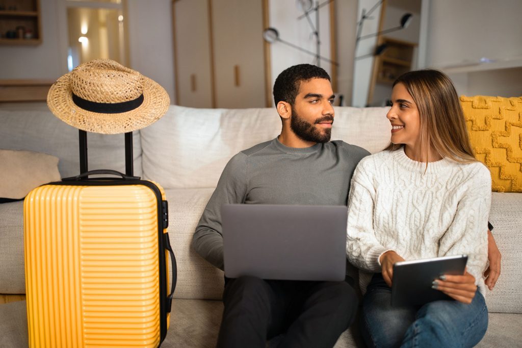 A couple sitting on a couch smiling at each other with a suitcase beside them